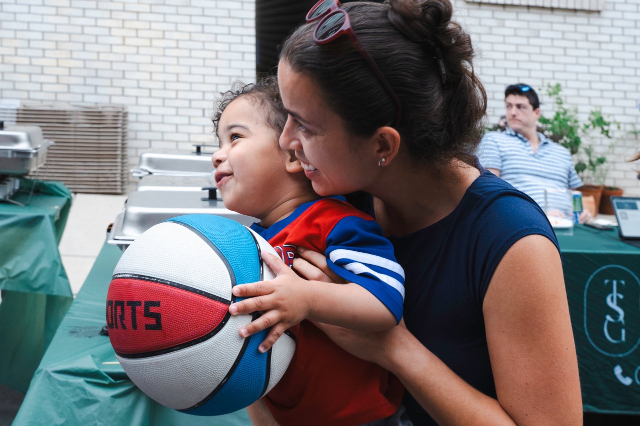 Woman helps a smiling child hold a colorful basketball at a neighborhood gathering.