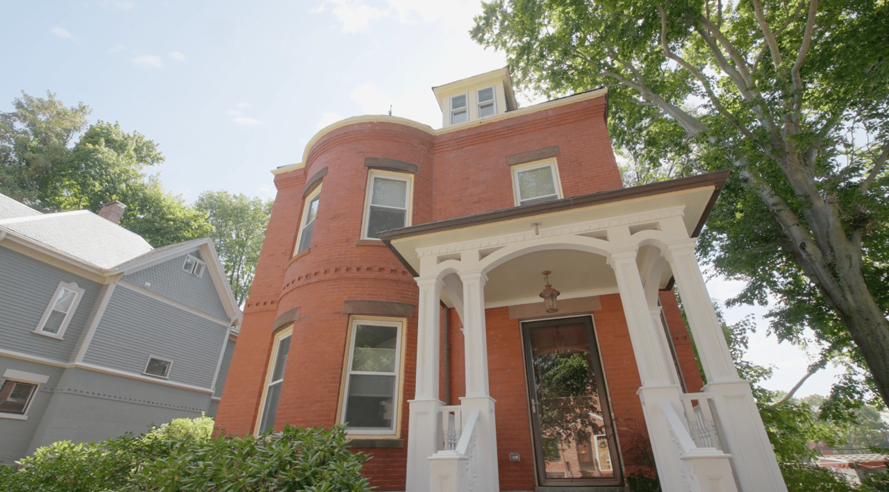 Red-brick home with a rounded bay and ornate white-columned porch beneath leafy trees.