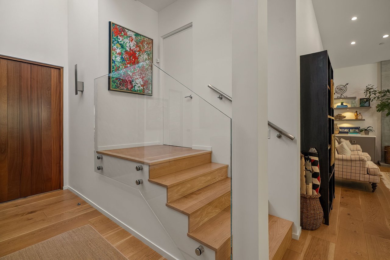Modern entry hall with wood stairs, glass railing, and colorful abstract artwork.