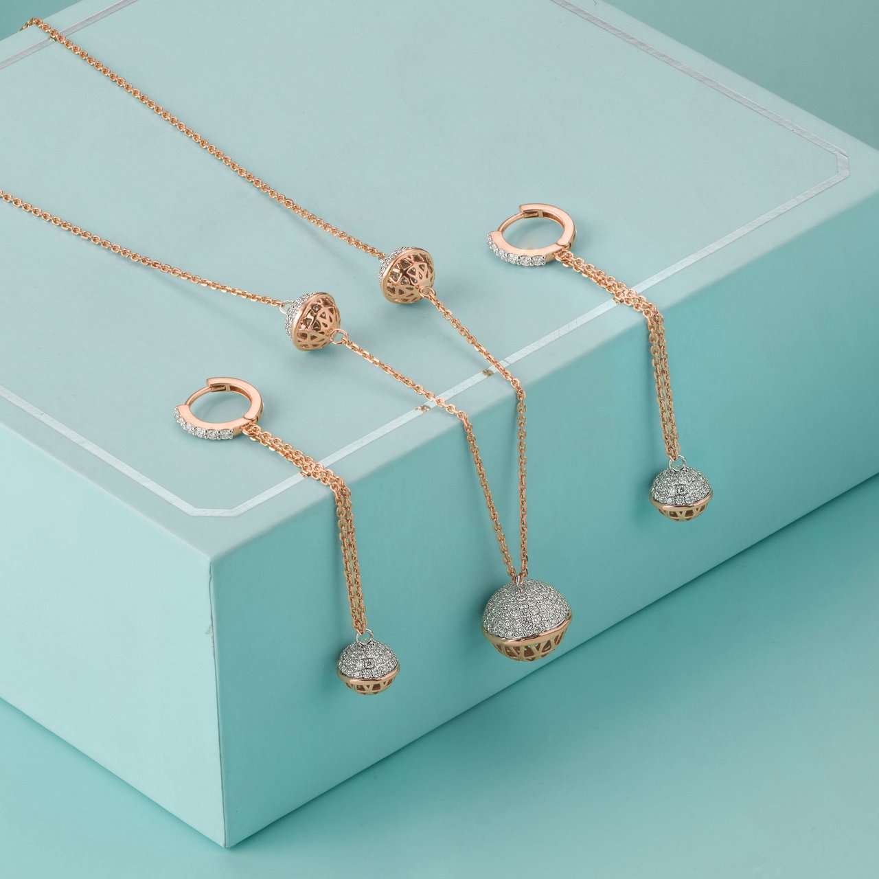 Rose gold necklaces and hoop earrings featuring pavé diamond ball pendants, draped over a teal display box on a matching teal background