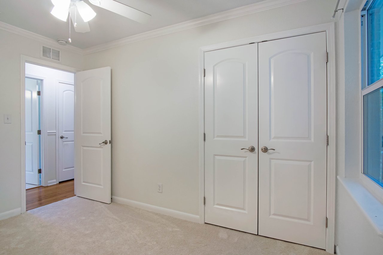 Empty room with beige carpet and cream walls. A ceiling fan with light is on. Open door leads to hallway. Closed double closet doors visible. Calm atmosphere.