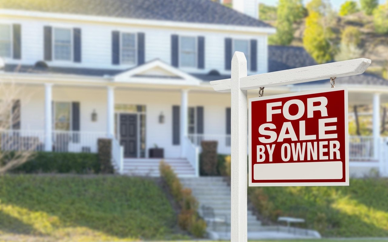 White two-story house behind a prominent red “For Sale by Owner” sign.