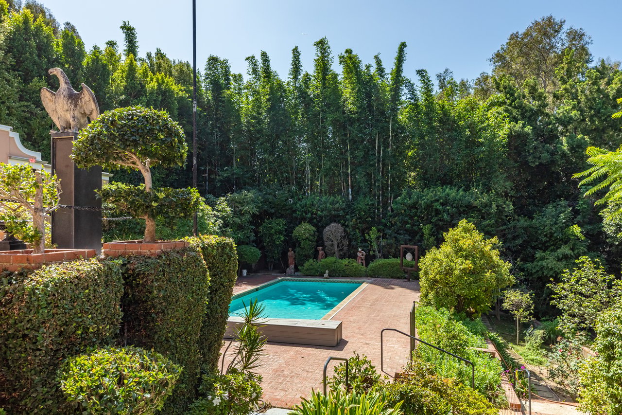 Backyard pool surrounded by a brick patio, sculpted hedges, and dense greenery.