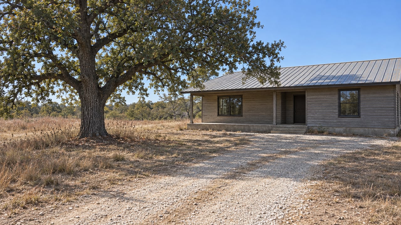 Low rural home with a metal roof beside a broad post oak, with a gravel drive across open ground.
