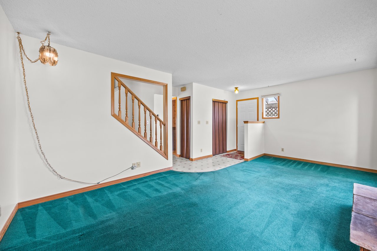 Mostly empty living room with turquoise carpet, a wood-railed staircase, and an entry hall.