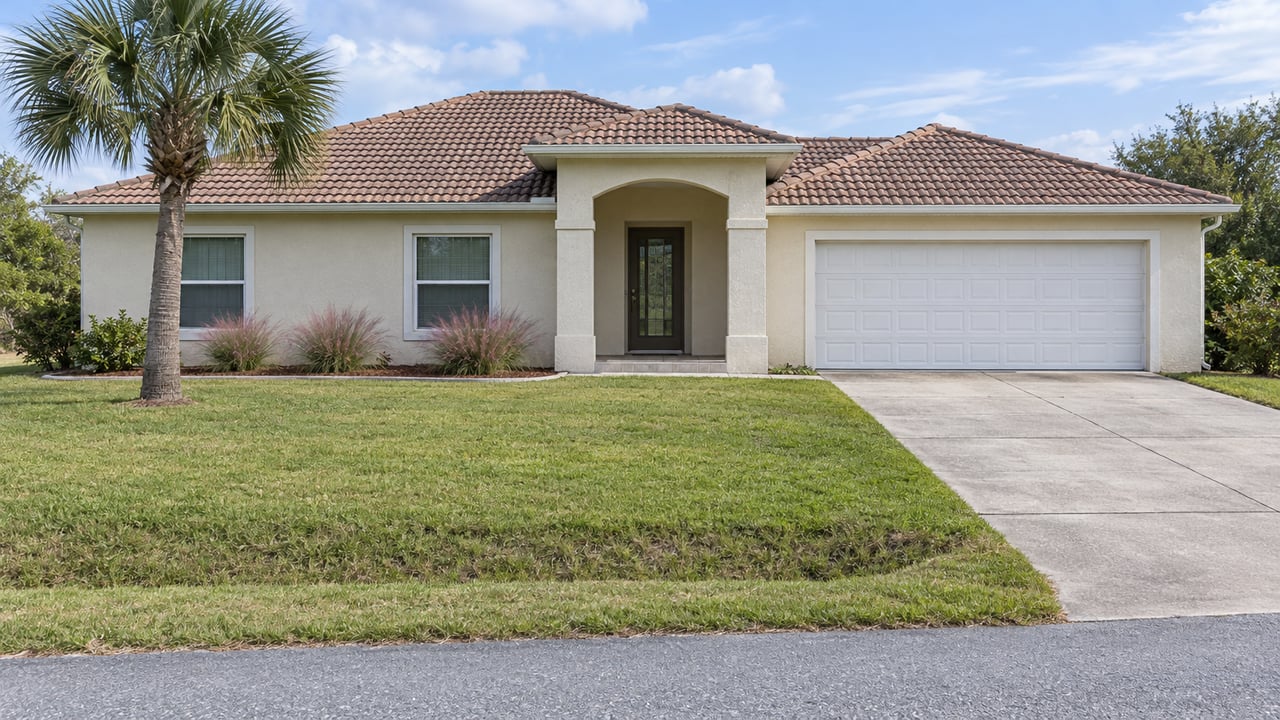 Cream-stucco single-story home beyond a broad lawn and shallow roadside swale, with a curved palm beside its arched entry.