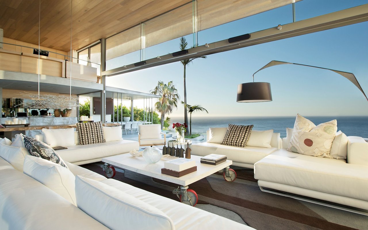 Modern ocean-view living room with white sectional sofas, floor-to-ceiling glass, and a wood ceiling.