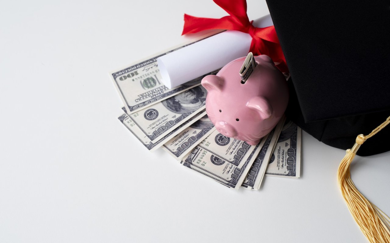 Pink piggy bank on dollar bills beside a diploma and graduation cap.