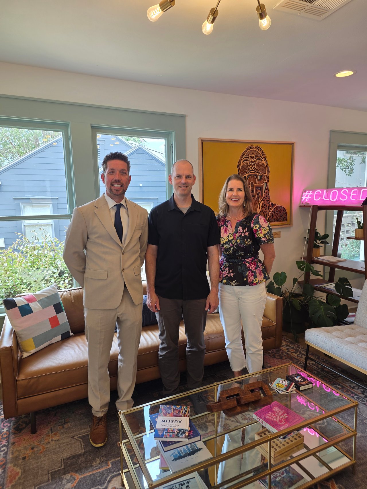 Three adults pose in a living room beside a neon “#CLOSED” sign.