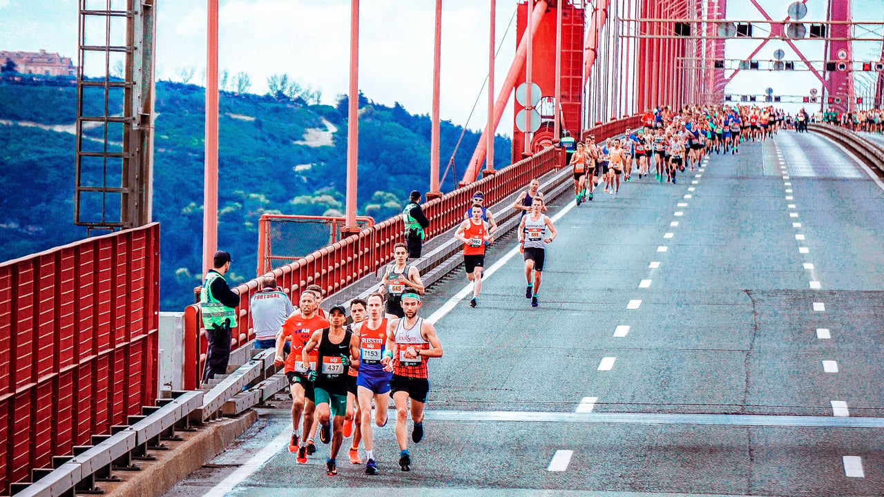 A panoramic photograph of thousands of marathon runners crossing the Verrazzano-Narrows Bridge at the start of the New York City Marathon, with New York Harbor and the Brooklyn skyline visible in the background.