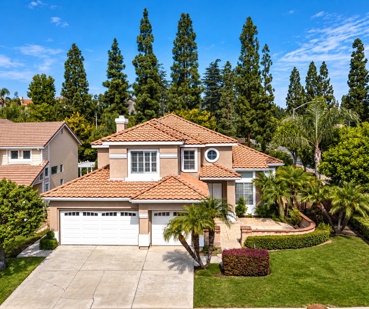 Two-story beige stucco home with a terracotta tile roof, circular window, and landscaped front yard.
