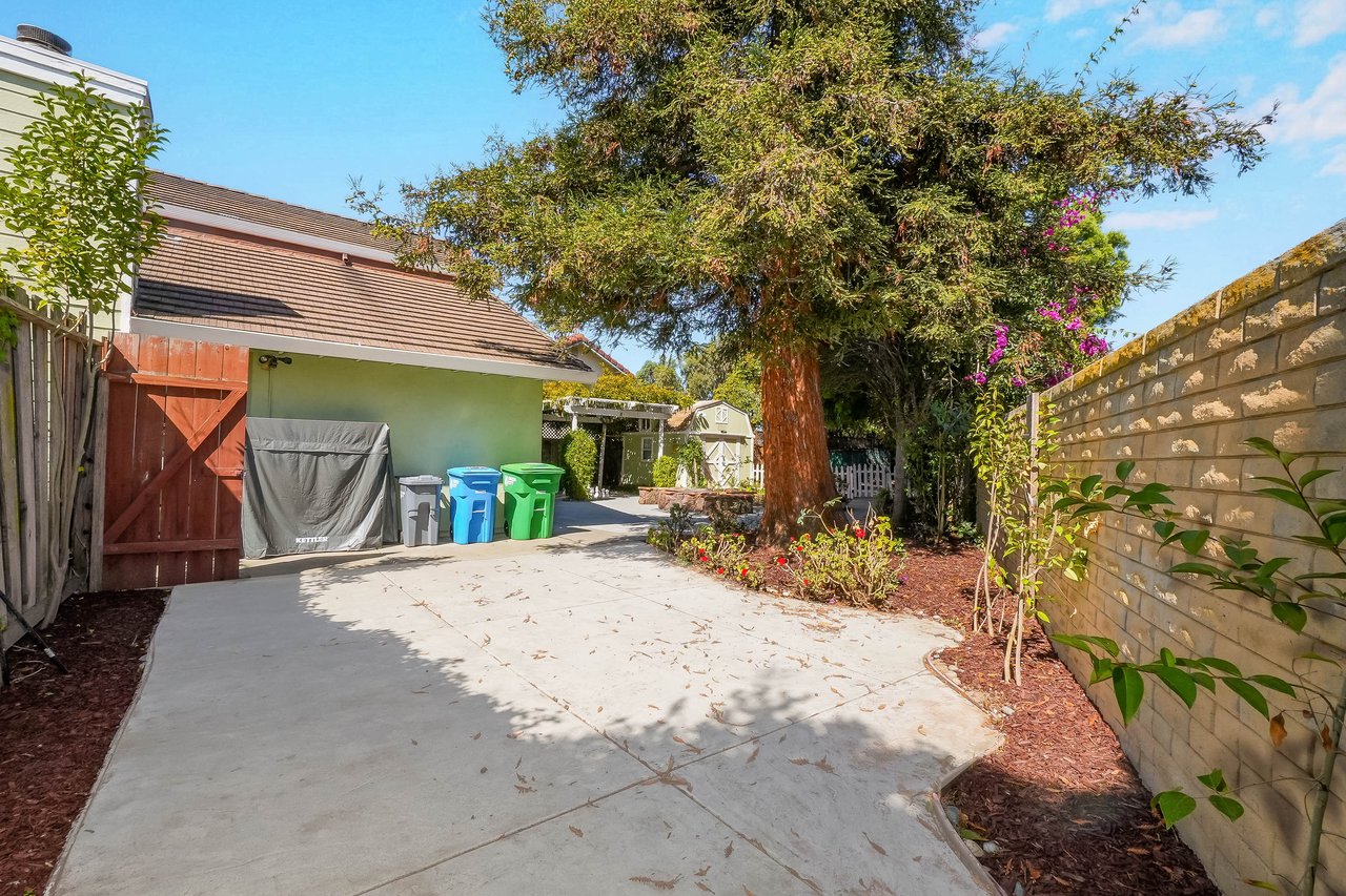 Paved backyard patio beneath a large tree, with a shed and stone wall.