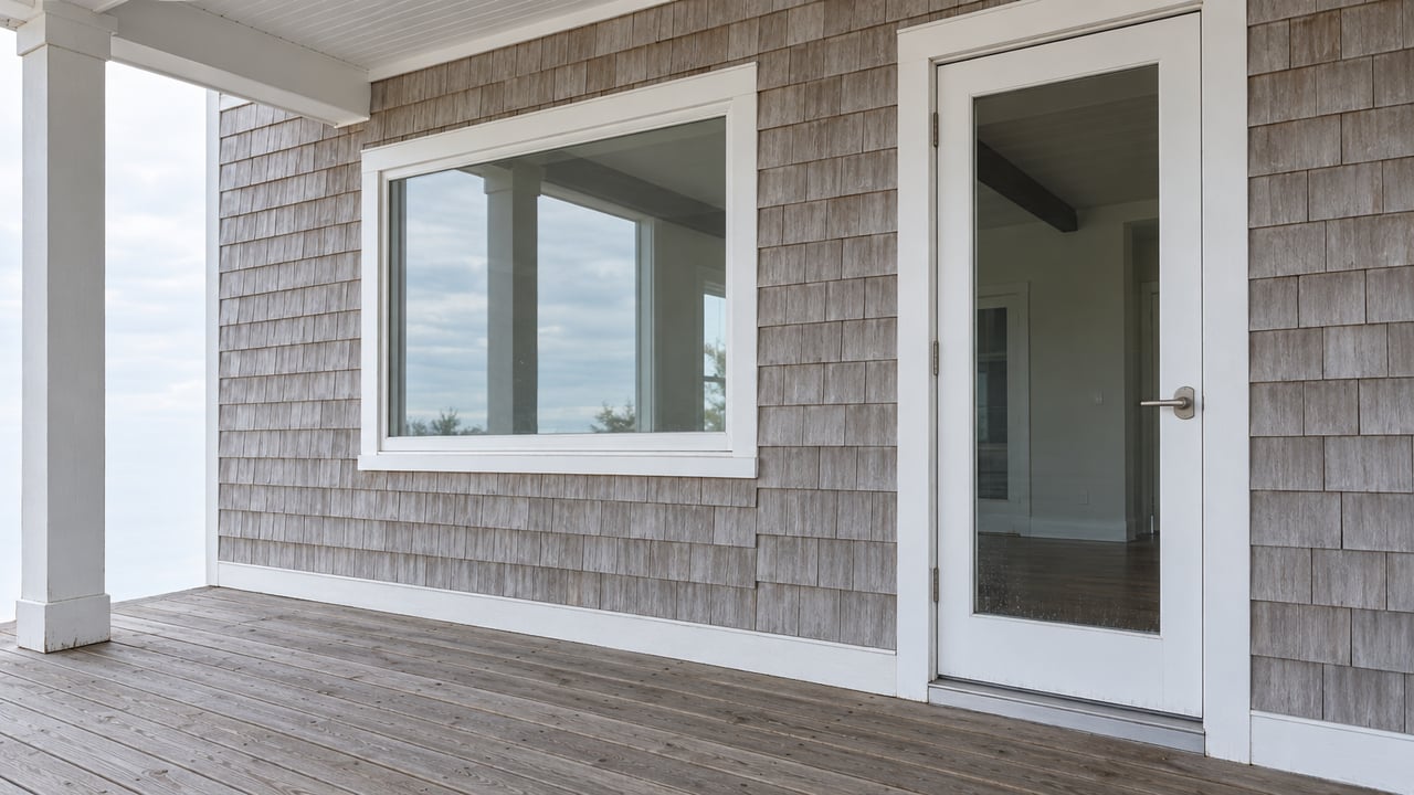 Weathered shingle siding, a closed glazed door, and a broad window above a gray wood porch deck in soft daylight.