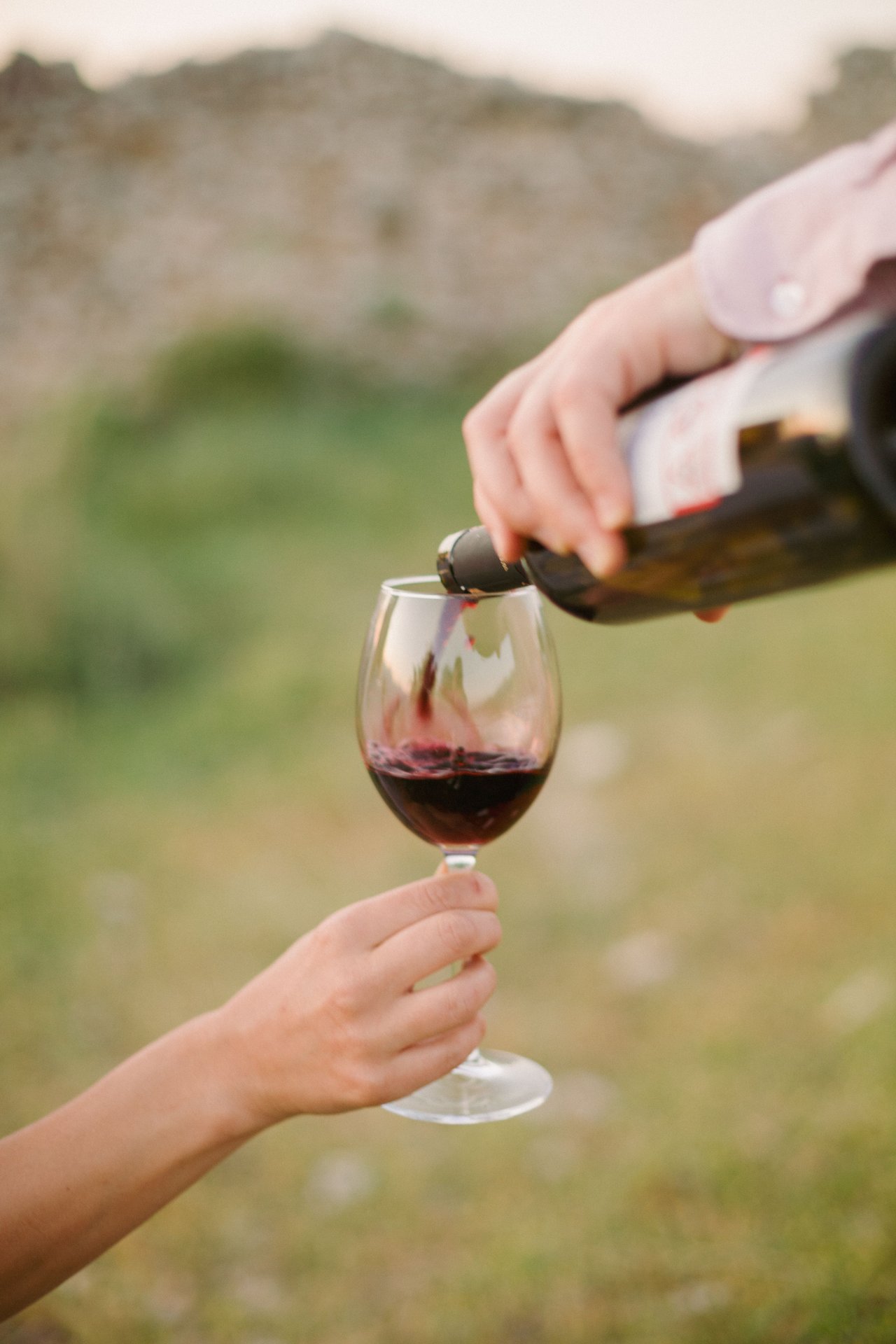 Red wine is poured into a glass held outdoors against a softly blurred green landscape.