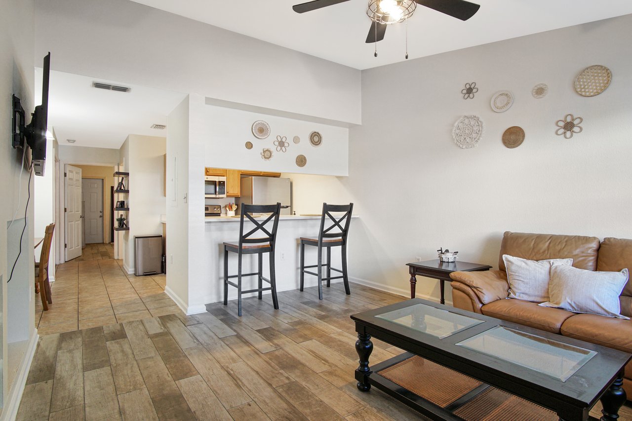 Cozy living room with tan leather sofa, glass coffee table, and cushion. Wall art and ceiling fan add charm. Open kitchen with bar stools visible.