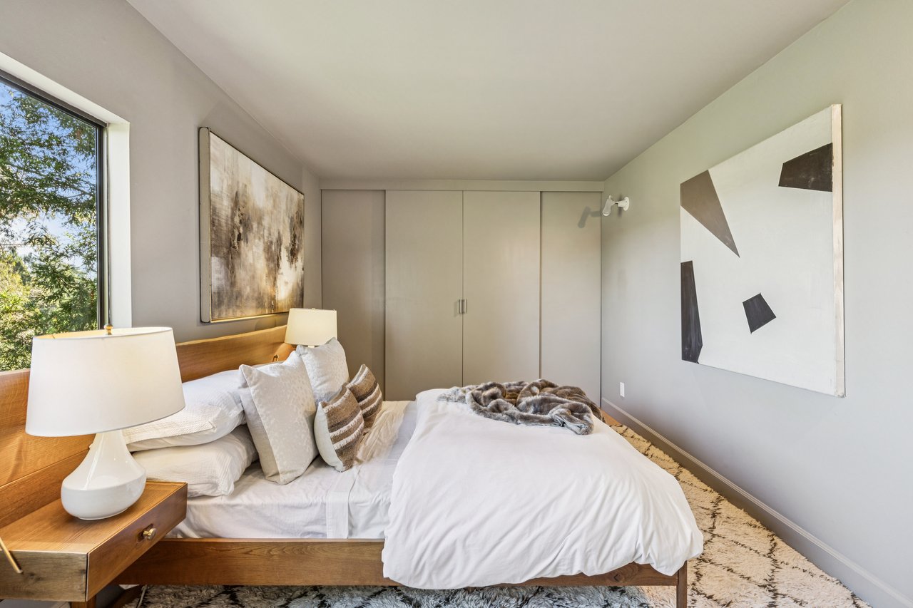 Bedroom with a wood platform bed, abstract art, and a large window overlooking trees.