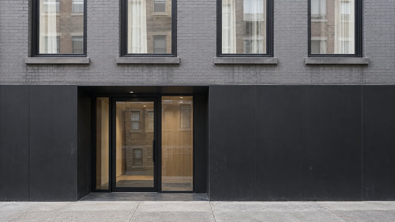 Gray masonry residential facade with black-framed windows, a recessed glazed entrance and a weathered concrete sidewalk.