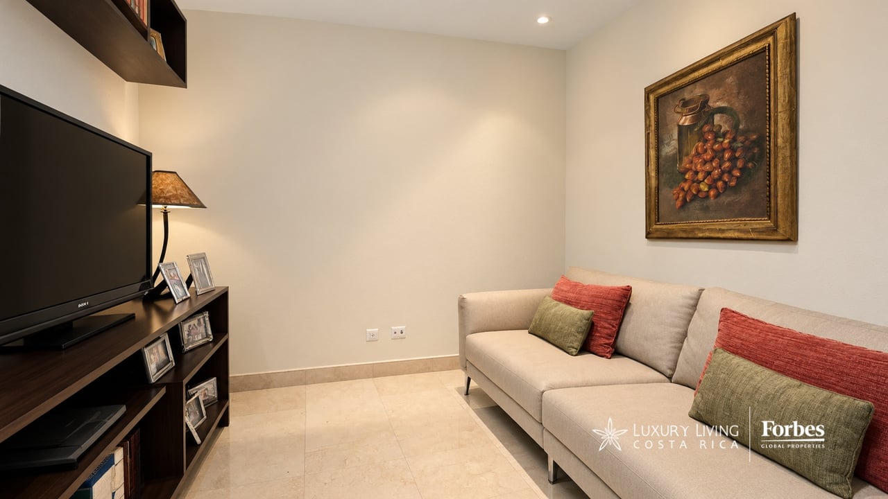 Furnished TV room with beige sectional sofa, dark media console, and framed fruit painting.