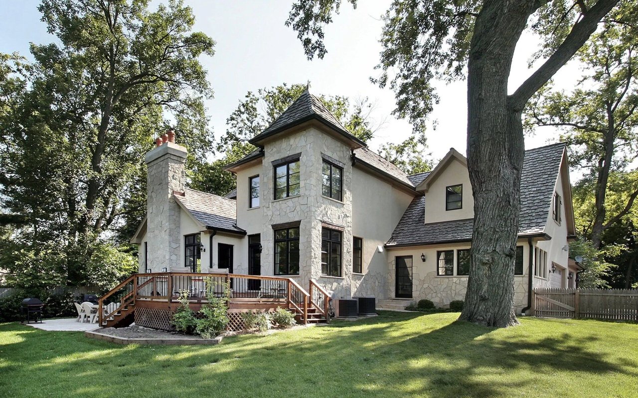 Two-story stone-and-stucco house with a raised deck, broad lawn, and mature shade trees.