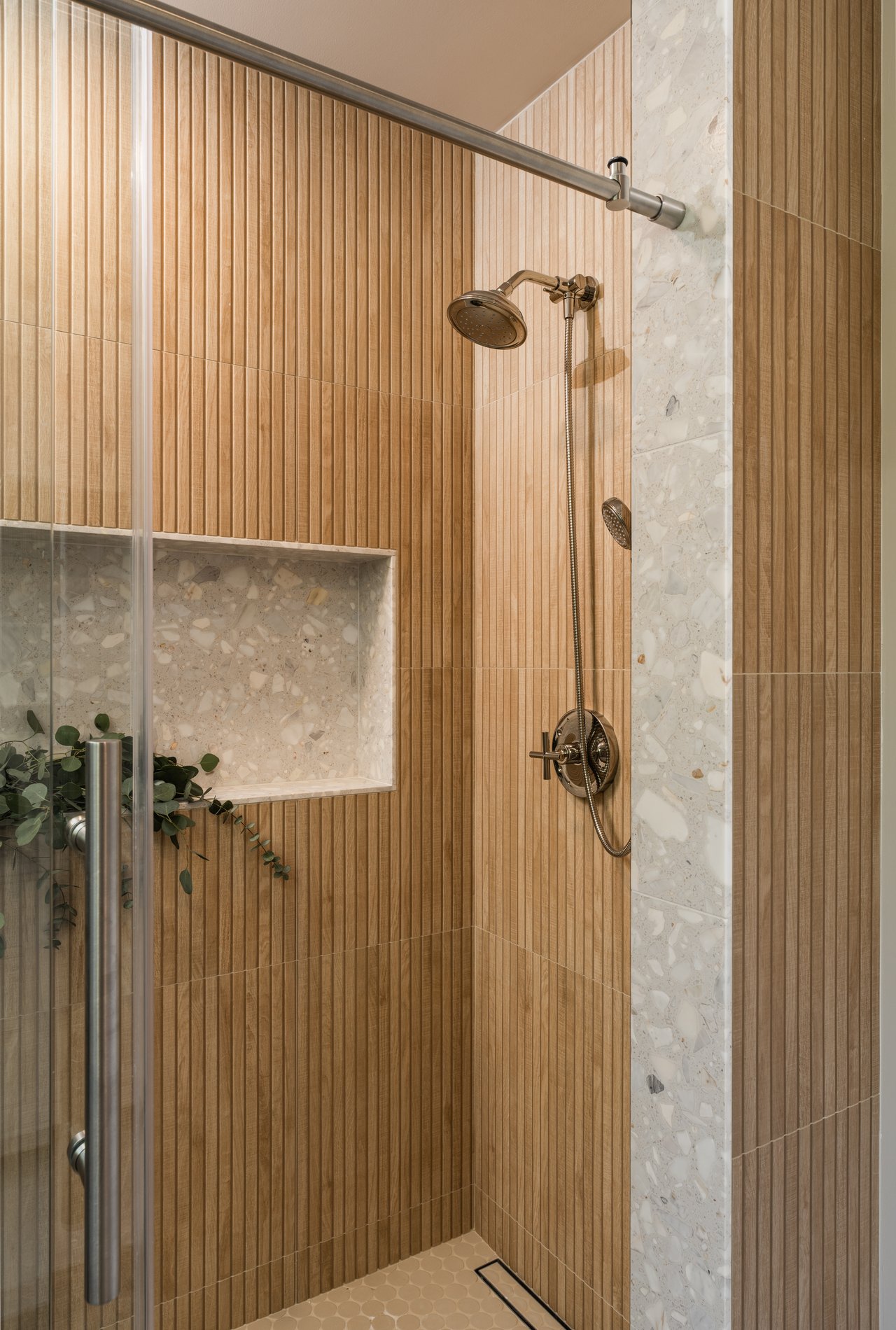 Walk-in shower with vertical wood-look tile, terrazzo panels, and bronze-toned fixtures.
