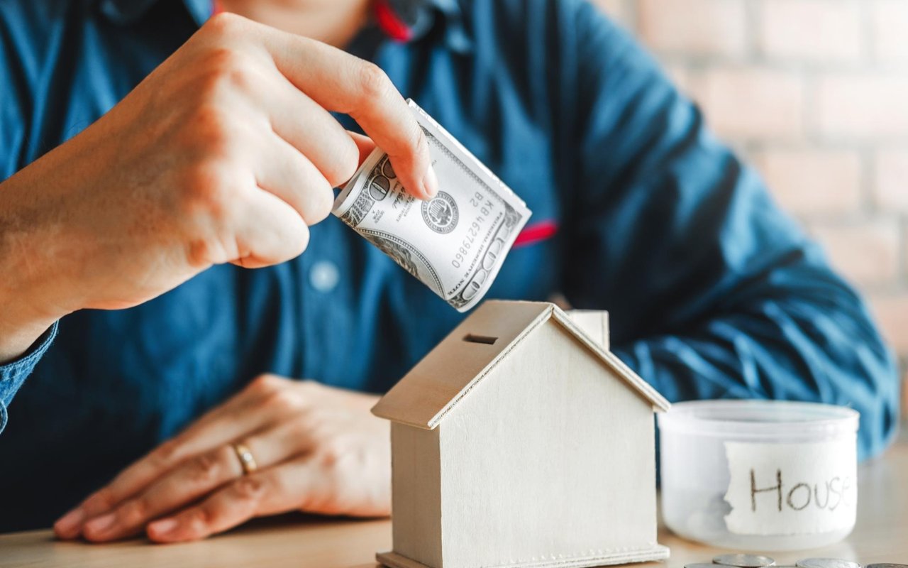 A person puts a dollar bill into a model house savings box beside a jar labeled “House.”