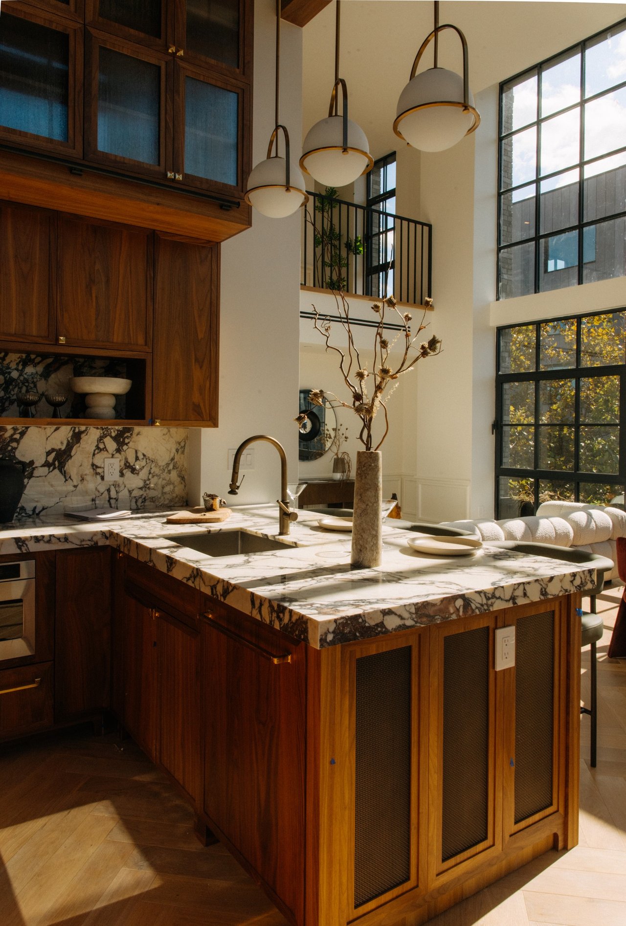 Kitchen with a marble island, walnut cabinetry, brass pendant lights, and tall black-framed windows.