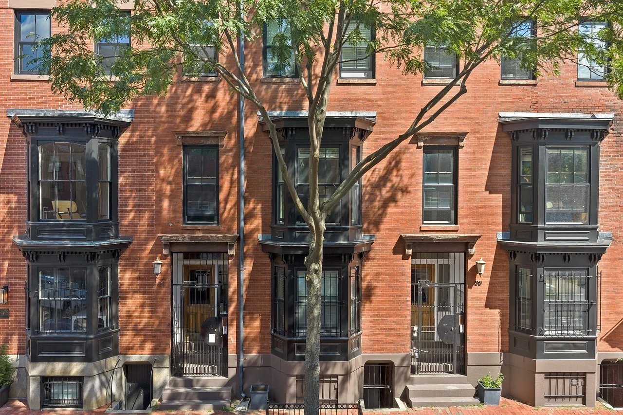 Red-brick townhouses with dark bay windows and iron entry gates beneath a leafy tree.
