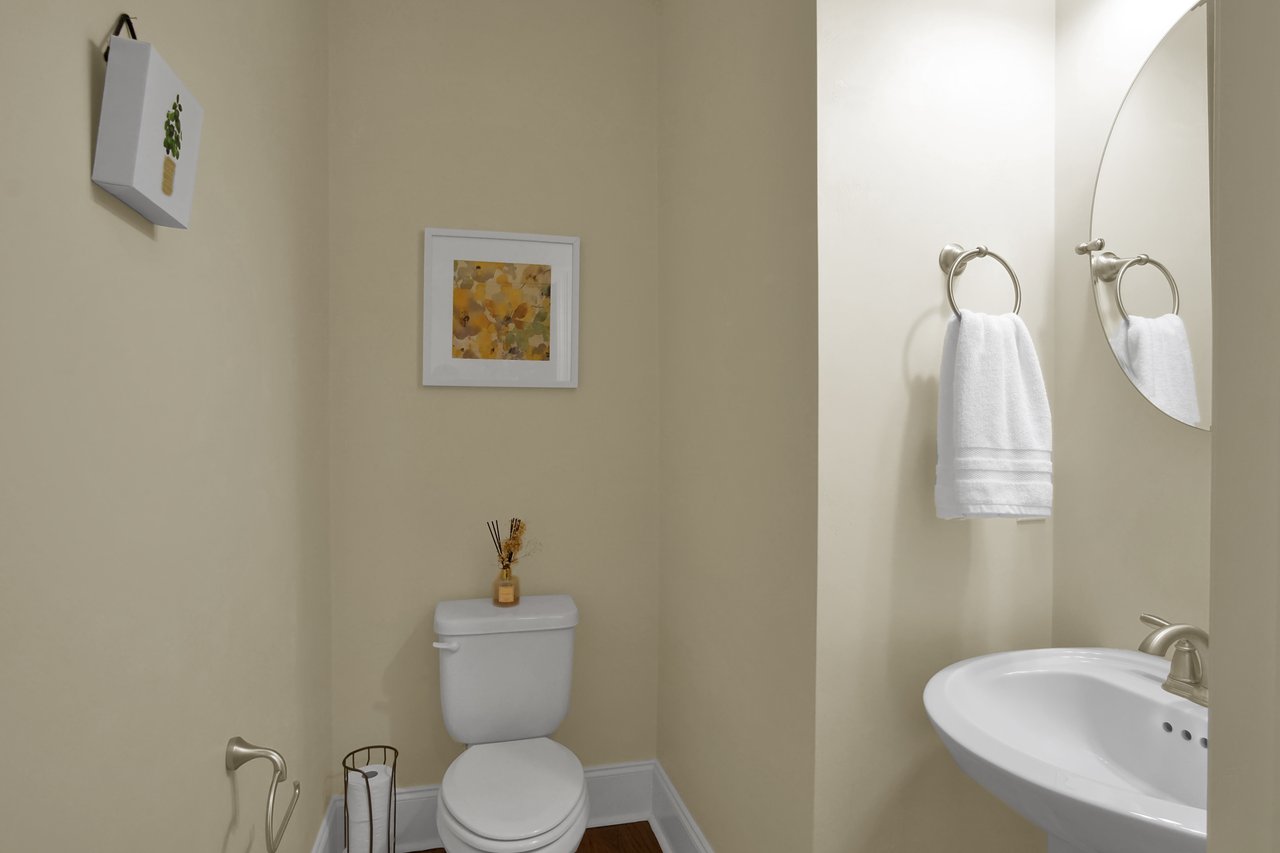 A small bathroom with light beige walls features a white toilet with decor on its tank. A simple framed artwork and plant decor adorn the walls. A pedestal sink with a round mirror and a white towel adds to the clean, minimal look.