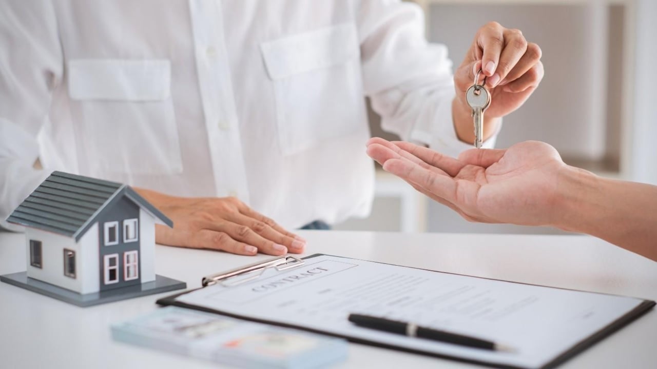 A key is handed over beside a miniature house and home sale contract.