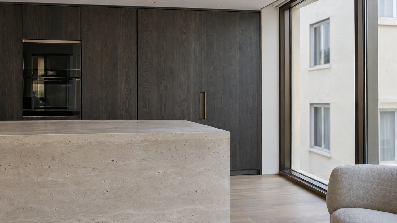 Limestone kitchen island beside a charcoal oak cabinet wall and floor-to-ceiling windows.