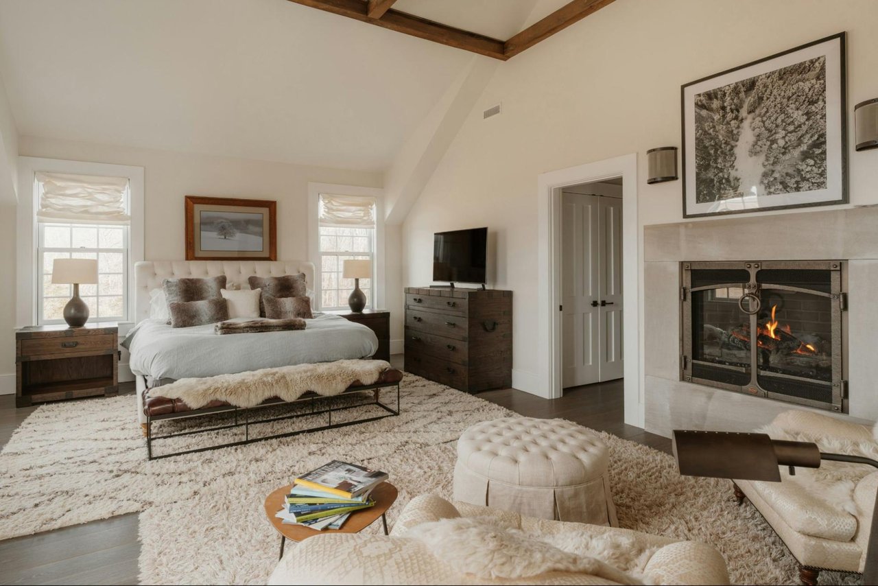 Staged luxury bedroom with a white bed, exposed wood beams, and a lit fireplace.