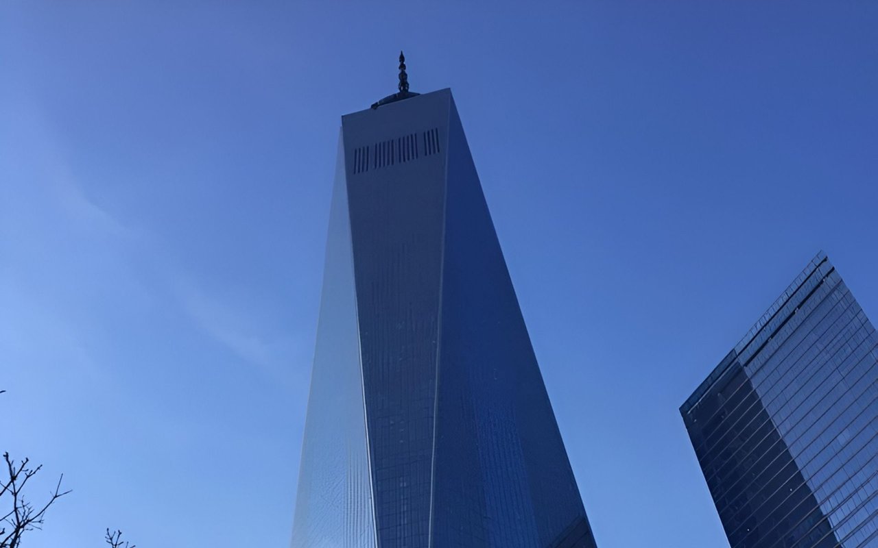 One World Trade Center rises above the frame against a blue sky, beside a glass skyscraper.