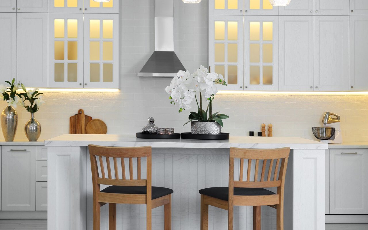 White kitchen with a marble-topped island, wooden chairs, and a white orchid centerpiece.