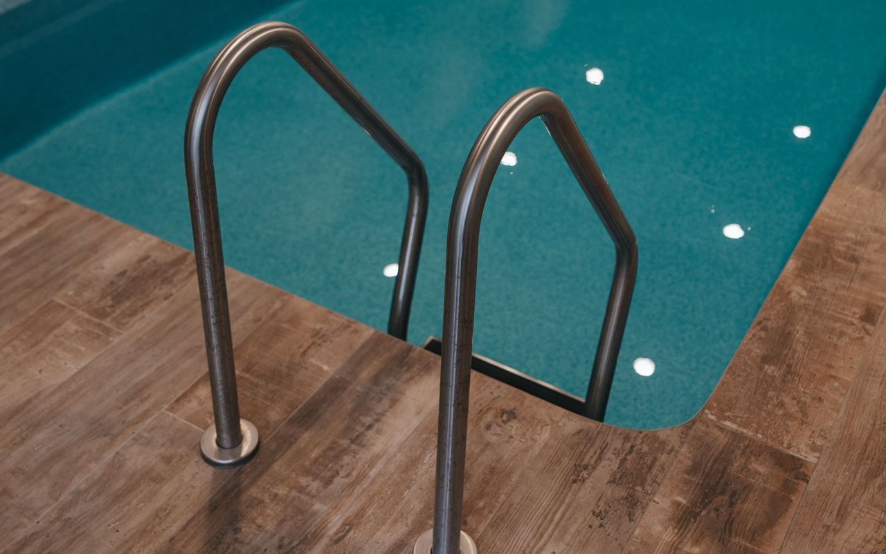 Indoor pool with turquoise water, a stainless-steel ladder, and wood-look tile decking.