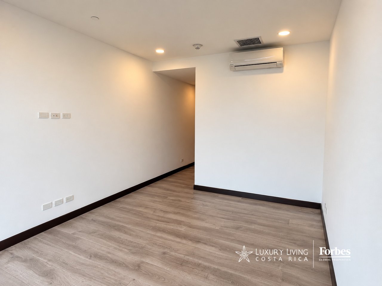Empty apartment room with wood-look flooring, recessed lights, and a wall-mounted air conditioner.