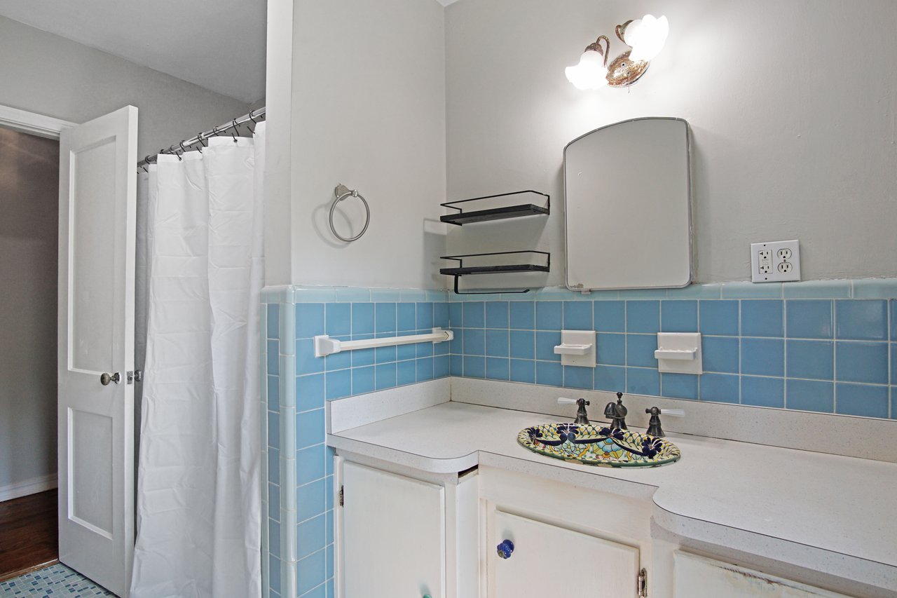 Vintage bathroom with light blue wall tiles, white counter, and a decorative basin. Open door and white shower curtain in the background, creating a serene tone.