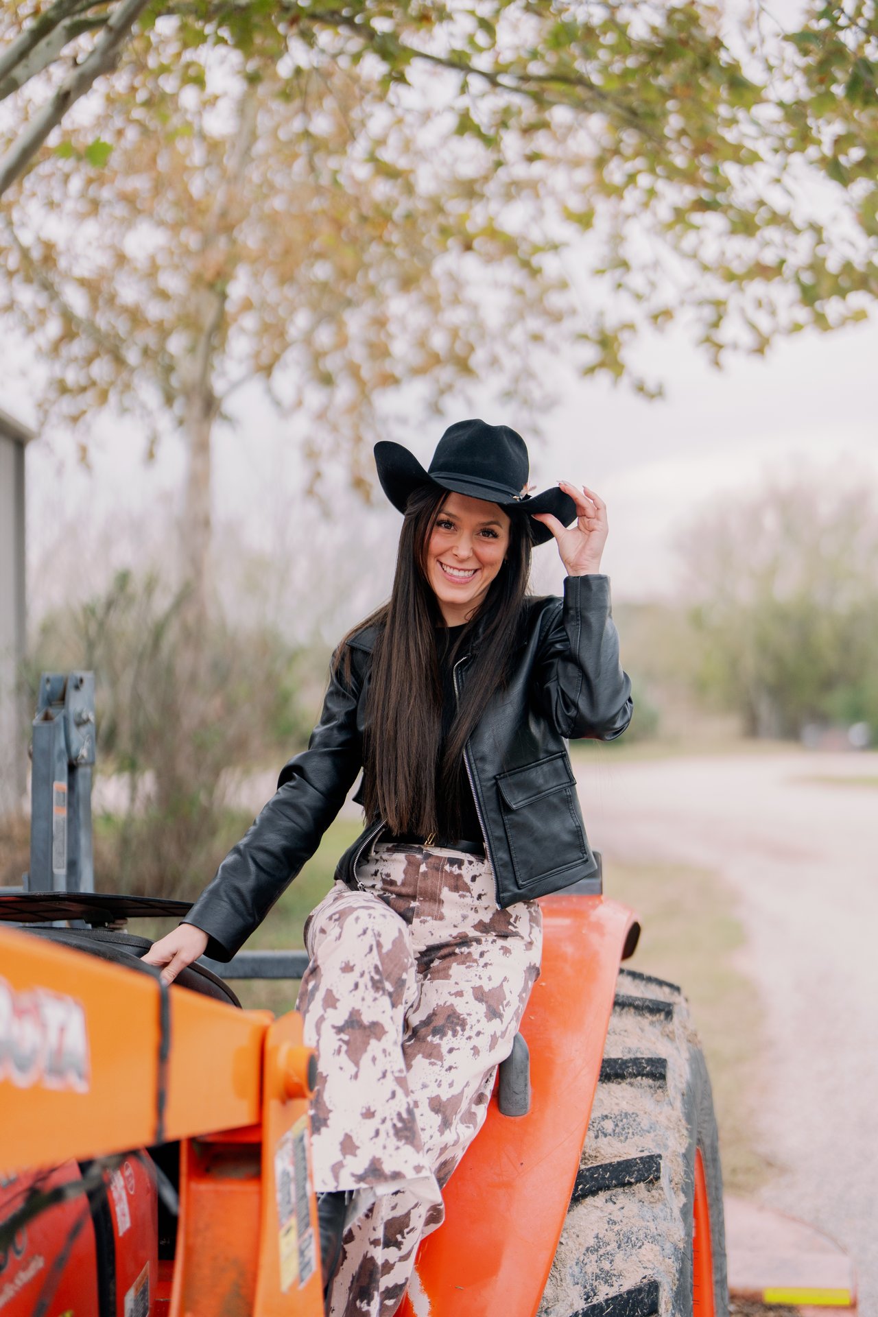 Smiling woman in a black cowboy hat and leather jacket seated on an orange tractor.