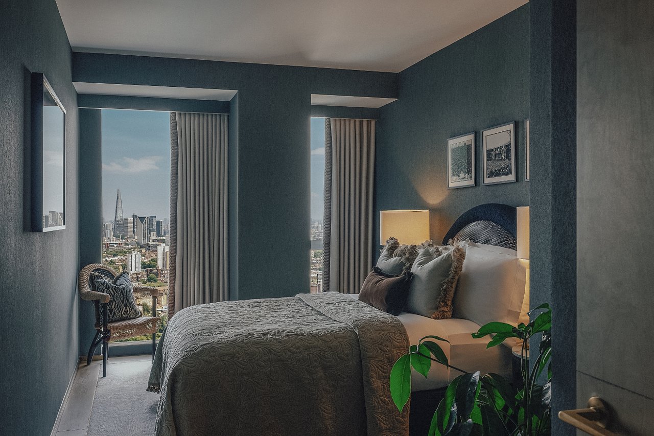 Bedroom with a large bed, dark teal walls, and London skyline views through tall windows.