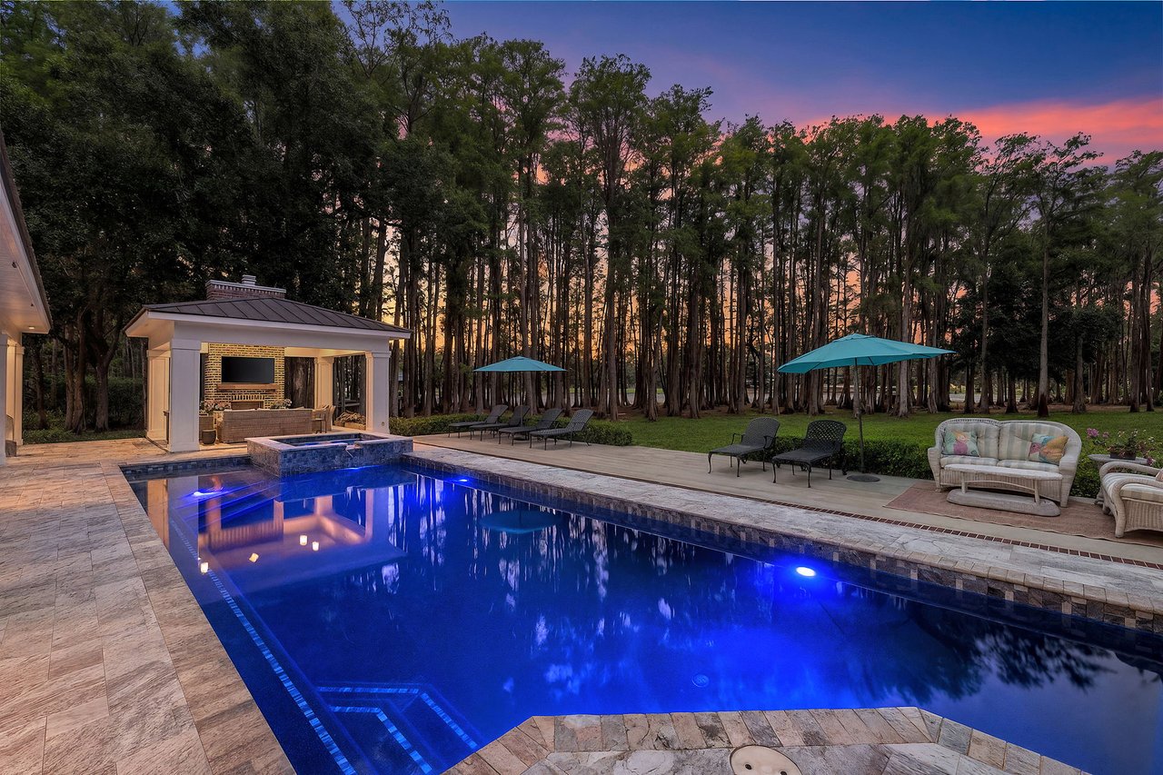 Illuminated backyard pool and stone patio with an outdoor kitchen and wooded backdrop at dusk.