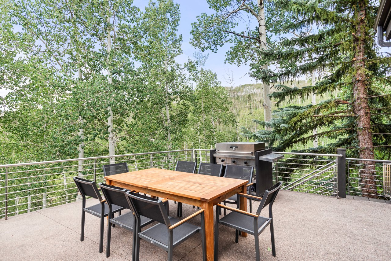 Furnished outdoor deck with a wooden dining table, built-in grill, and wooded hillside views.