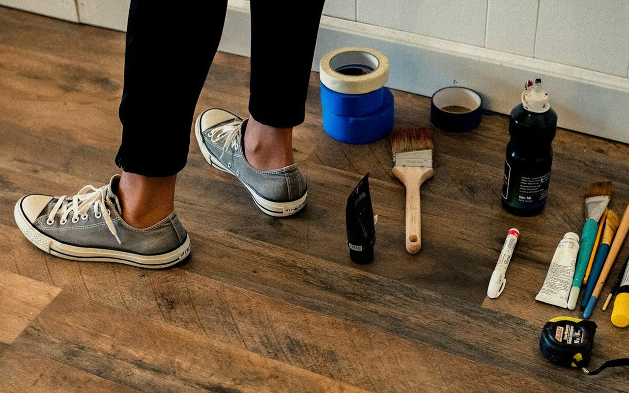 Painting supplies and a person’s sneakers on a wood floor during a home repair.