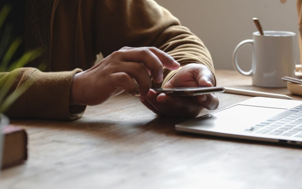 Person using a smartphone beside a laptop at a wooden table.
