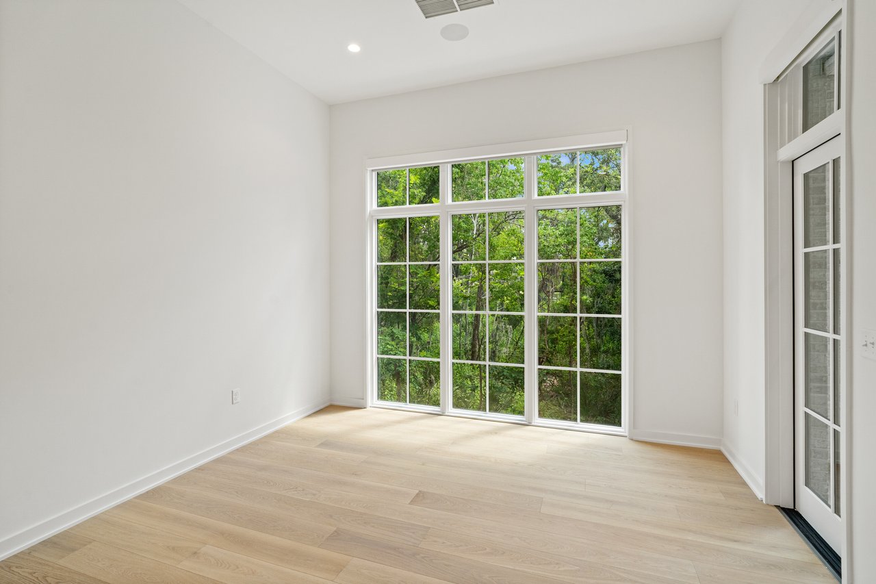Bright vacant room with pale wood floors and a large window overlooking trees.