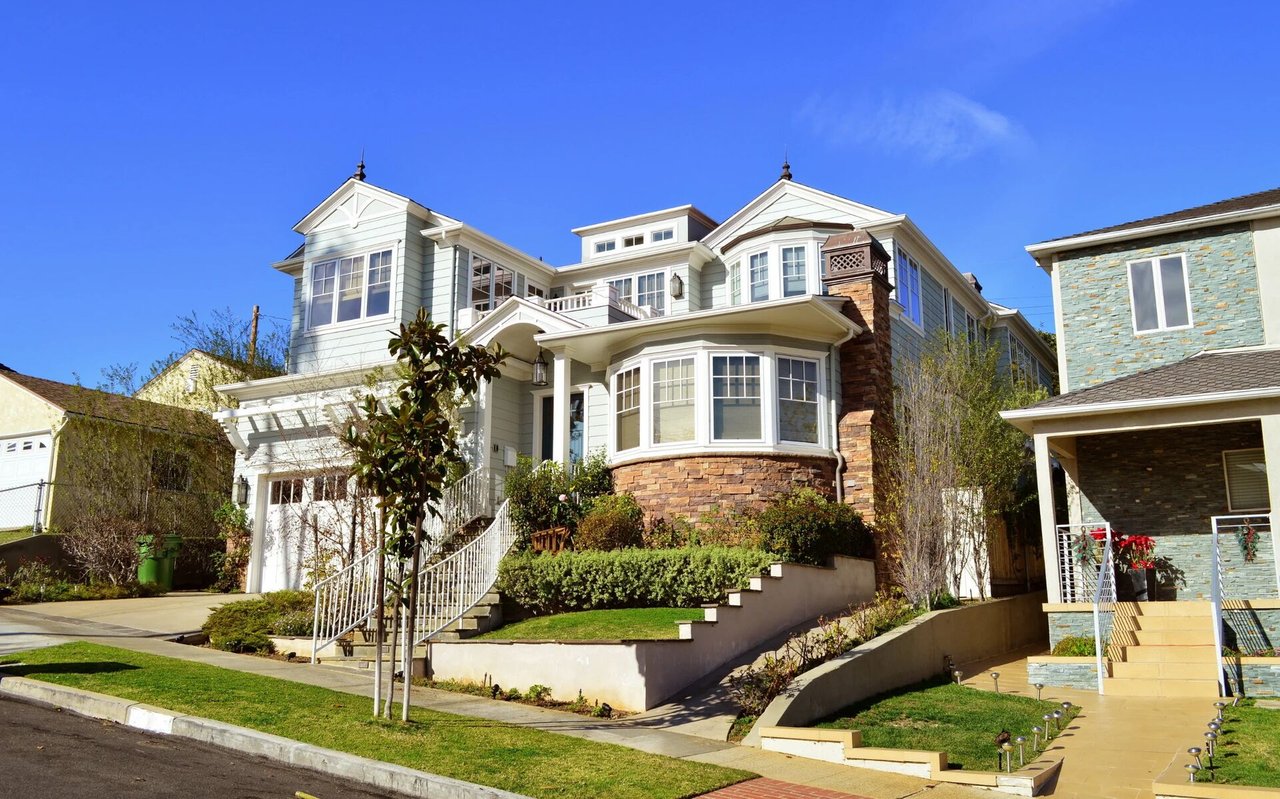 Large light-colored house with a stone-faced bay window, landscaped steps, and neighboring homes.