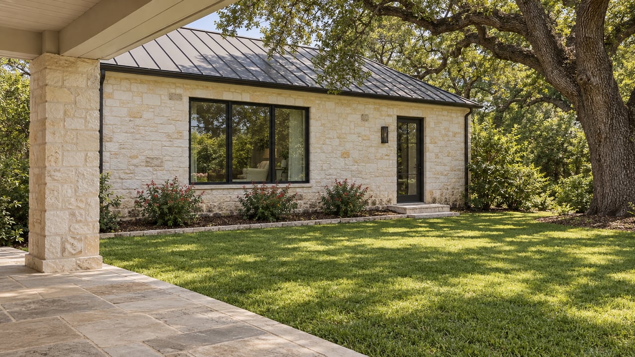A shaded limestone porch looks across a sunlit lawn toward a pale stone home with black-framed windows and a mature spreading oak.
