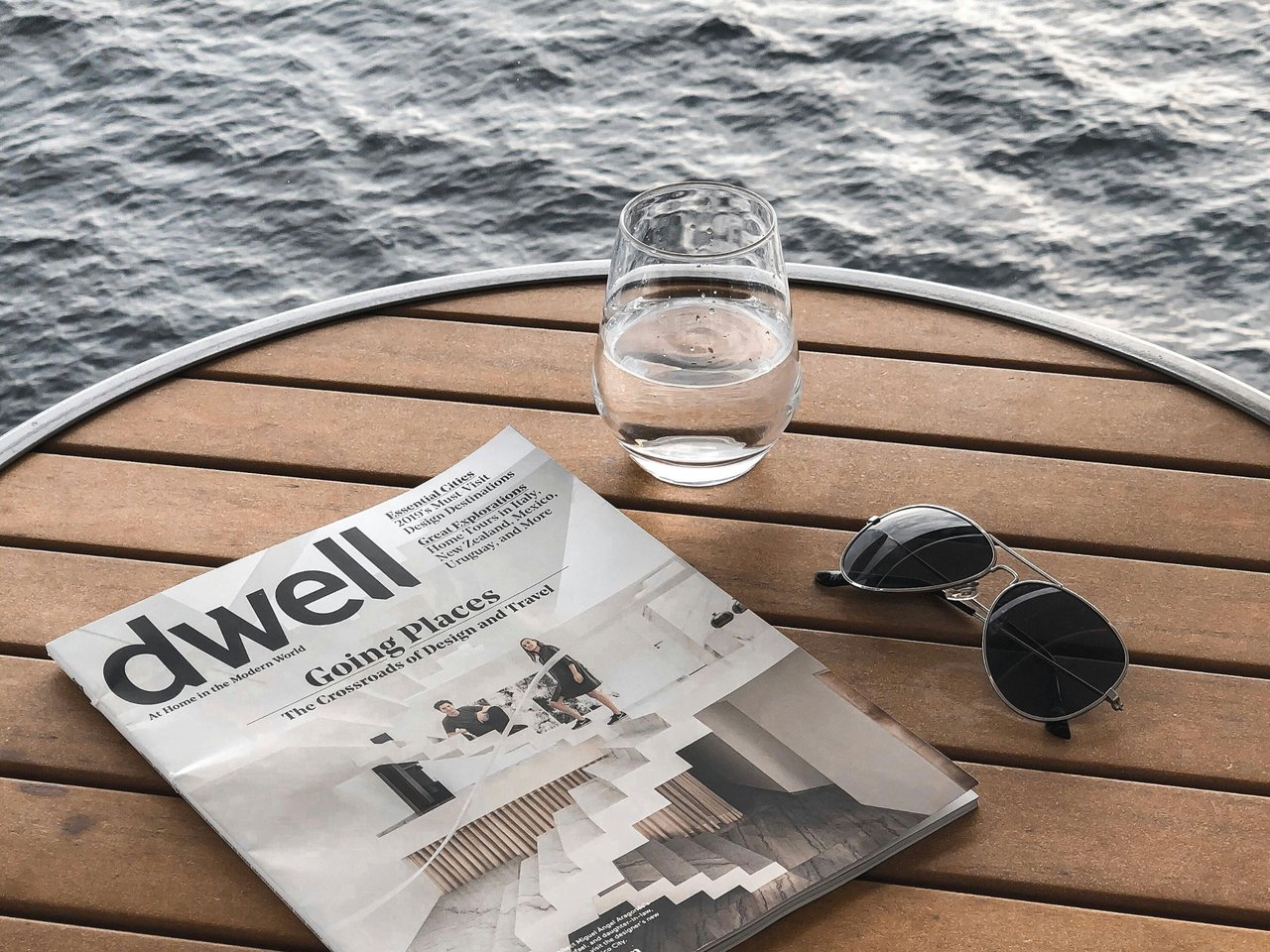 Dwell magazine, water glass, and aviator sunglasses on a wooden table beside the water.