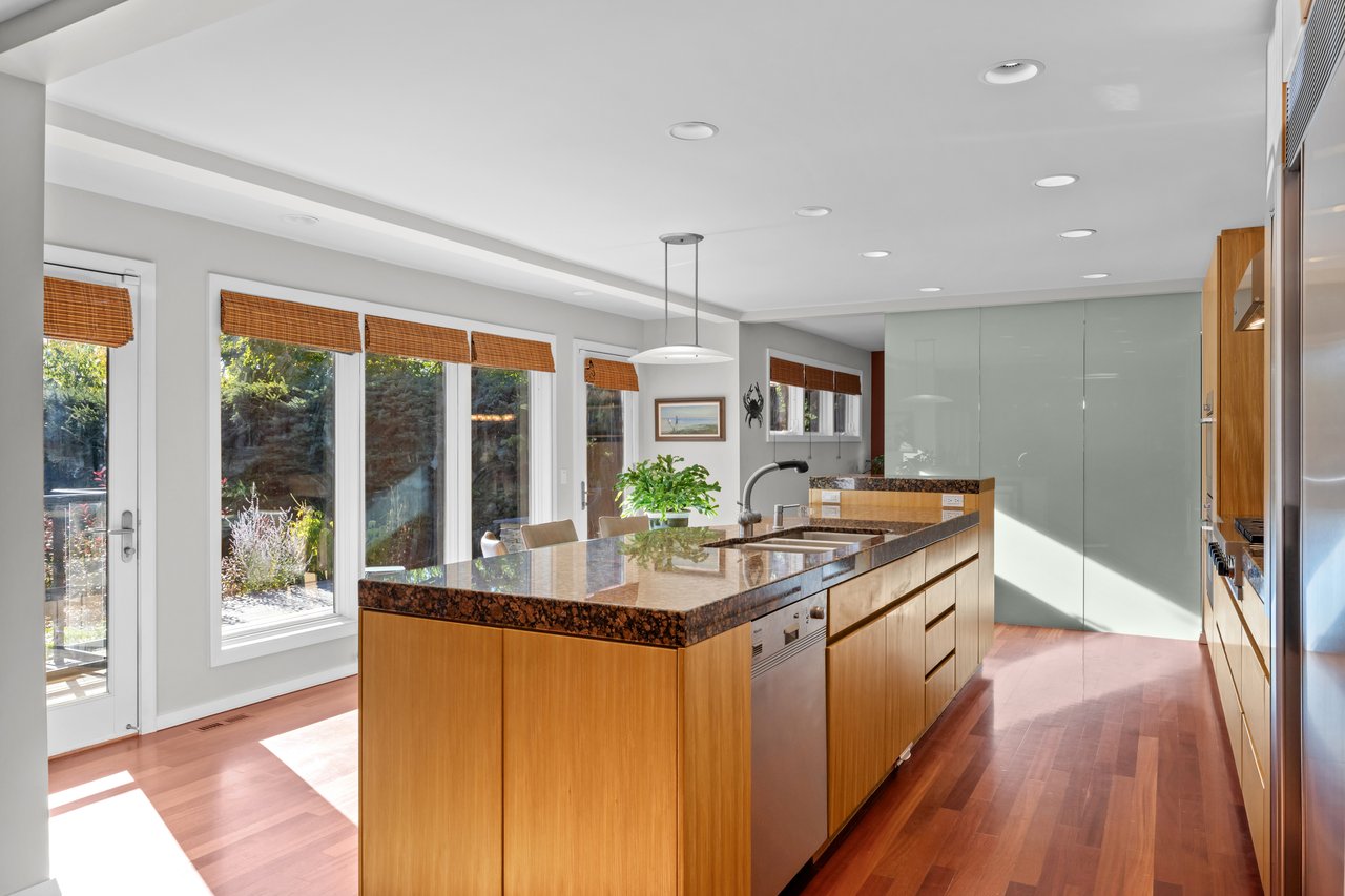 Modern kitchen with a long wood island, dark stone countertop, and large windows overlooking greenery.