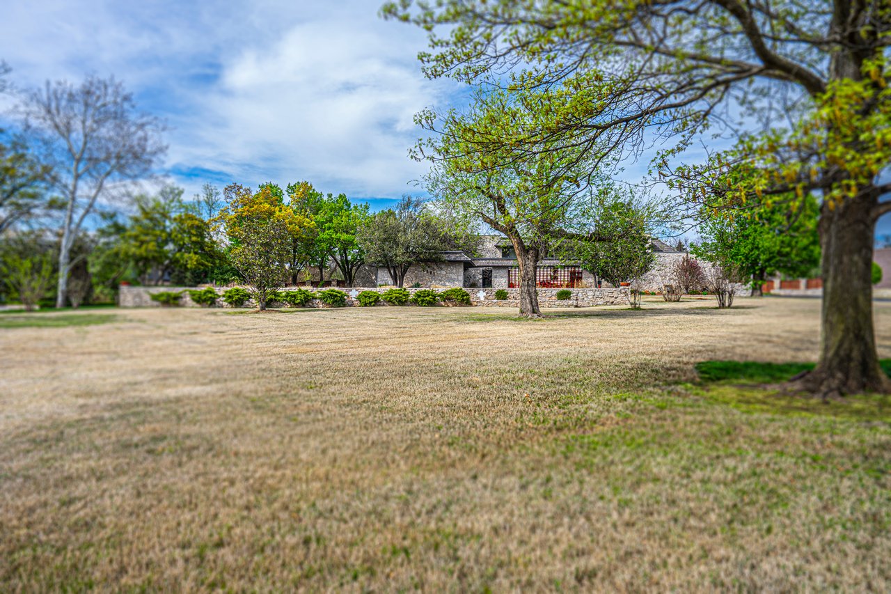 Single-story stone house beyond a broad lawn, framed by mature trees and a low stone wall.