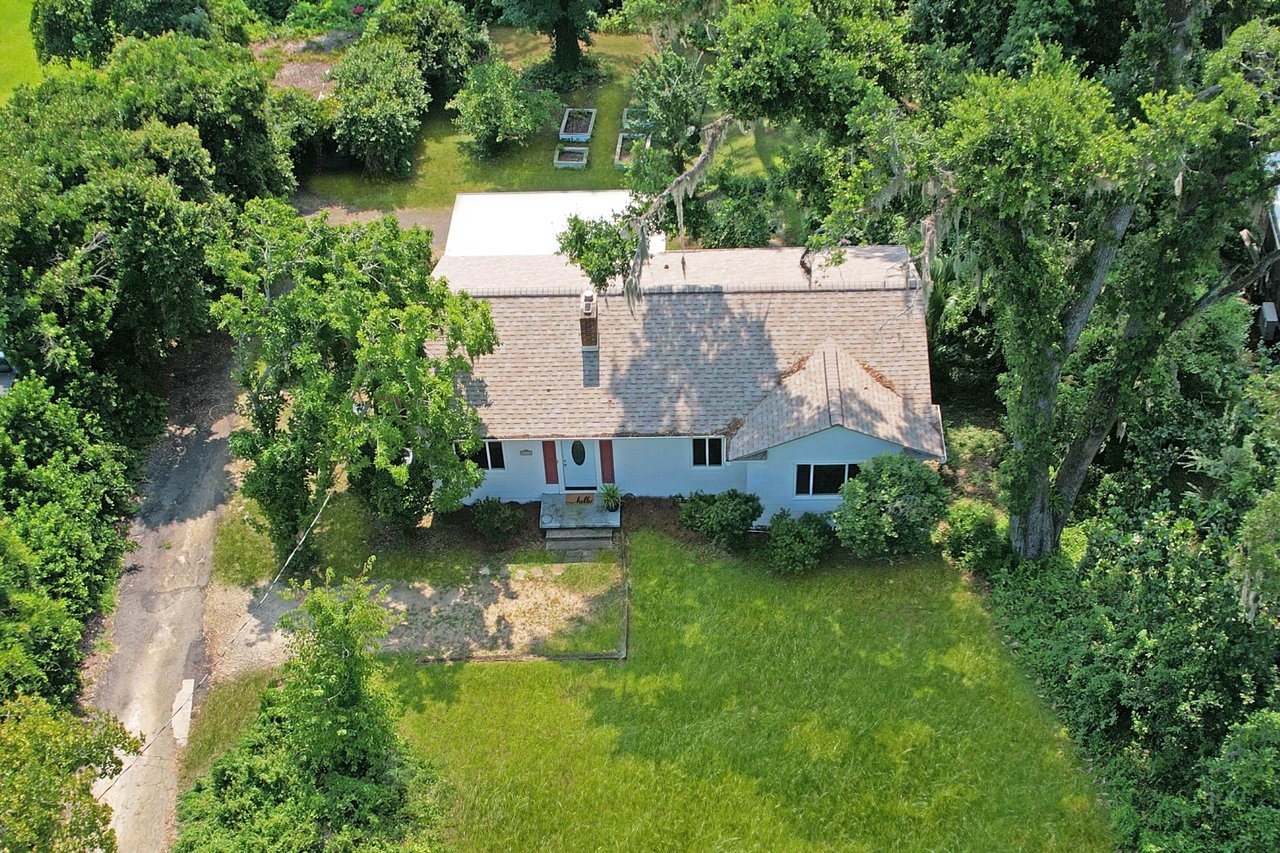 Aerial view of a small house with a light brown roof surrounded by lush green trees and grass. A pathway leads to the front porch, creating a serene and inviting atmosphere.