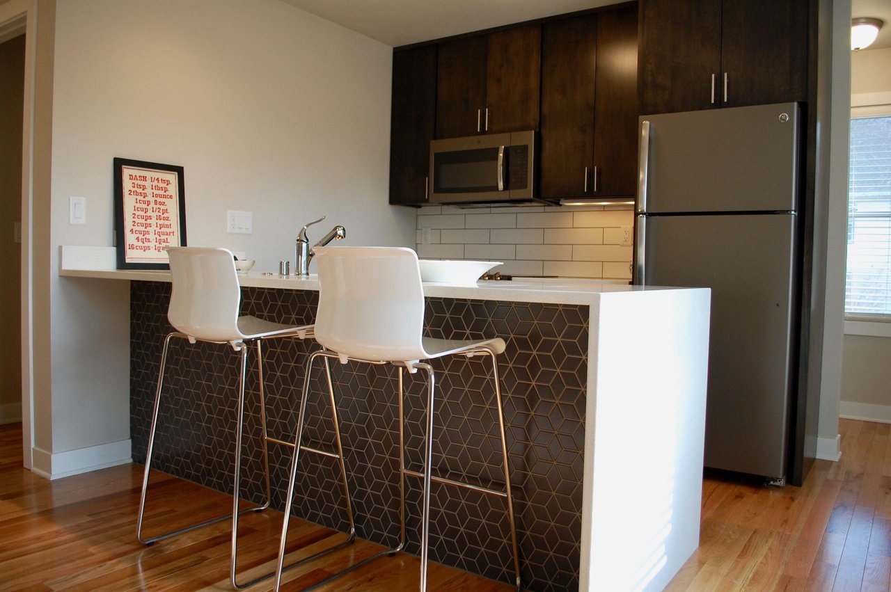 Apartment kitchen with dark cabinets, stainless steel appliances, and a breakfast bar with two white stools.